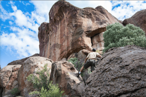 A person climbs a rocky formation under a blue sky with scattered clouds, surrounded by shrubs and boulders.