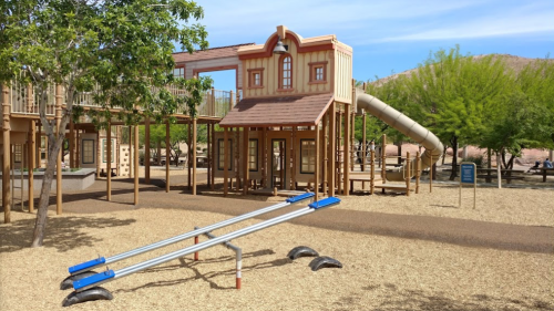 A playground with a western-themed structure, slide, and seesaw, surrounded by trees and gravel.
