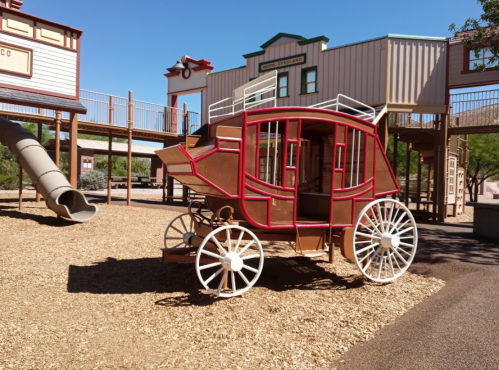 A wooden play structure resembling a stagecoach, set in a playground with a slide and western-themed buildings in the background.