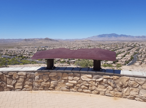 A scenic overlook with a metal plaque, featuring a view of a valley and distant mountains under a clear blue sky.