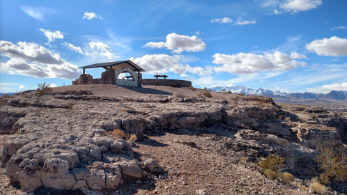 A rocky landscape with a shelter and picnic area under a blue sky with clouds, overlooking distant mountains.