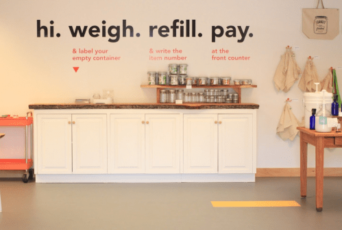 A minimalist store interior with a counter, jars, and a wall sign instructing customers on the refill process.