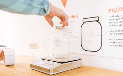 A person places a jar on a scale, with instructions for filling and weighing items displayed on the wall.