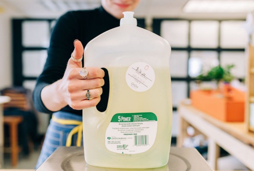 A person holds a large jug of liquid cleaner, showcasing the label and handle, in a bright indoor setting.