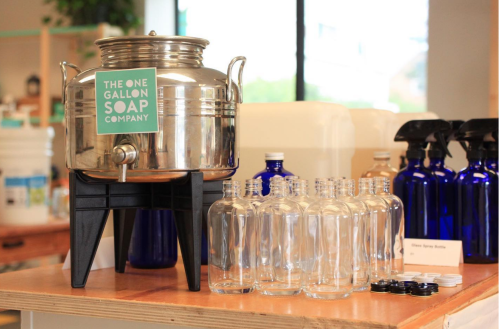 A silver soap dispenser on a stand with clear bottles arranged beside it in a bright, indoor setting.