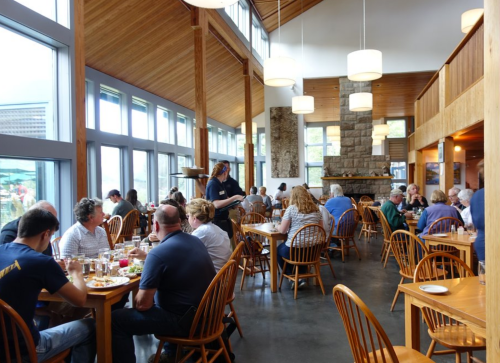A busy restaurant interior with wooden decor, large windows, and diners enjoying meals at tables.