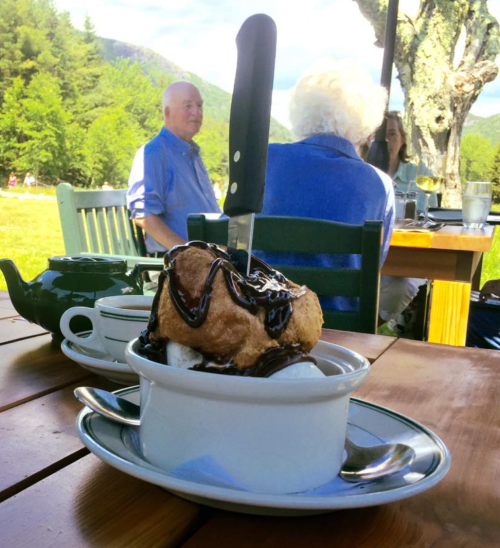 A dessert with ice cream and chocolate sauce sits on a table, with people enjoying a scenic outdoor setting in the background.