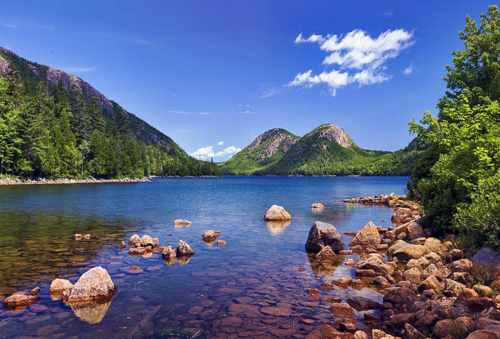 A serene lake surrounded by green mountains and rocky shores under a clear blue sky with scattered clouds.