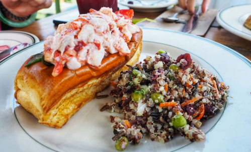 A lobster roll topped with fresh lobster meat, served alongside a colorful quinoa salad on a white plate.