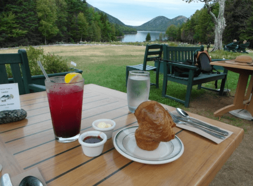 A table set outdoors with a drink, a roll, and dipping sauces, overlooking a scenic lake and mountains.