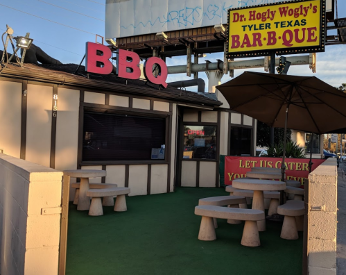 Exterior of a BBQ restaurant with a large sign, outdoor seating, and a shaded umbrella.