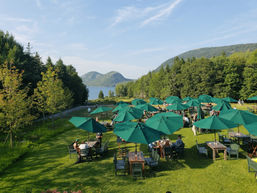 A scenic outdoor dining area with green umbrellas, surrounded by trees and mountains, overlooking a calm lake.