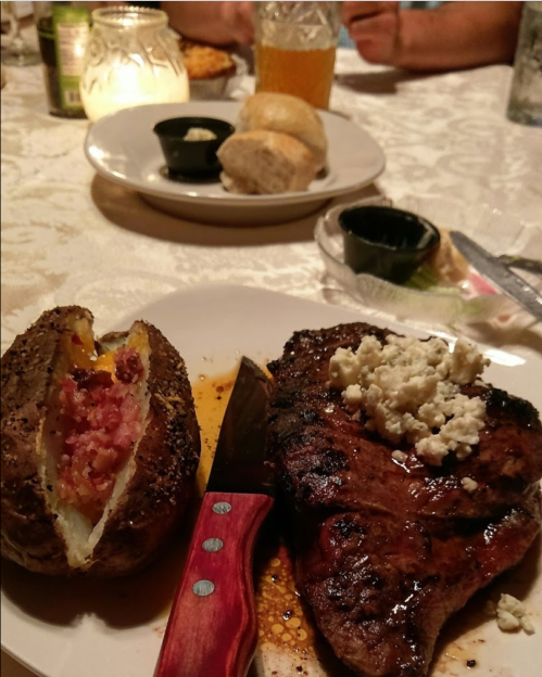 A plate with a grilled steak topped with cheese, a baked potato with bacon, and rolls in the background.
