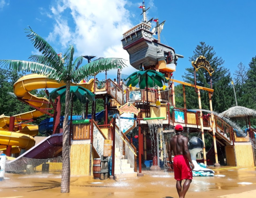 A water park with a pirate ship structure, slides, and palm trees; a person in red shorts walks nearby.