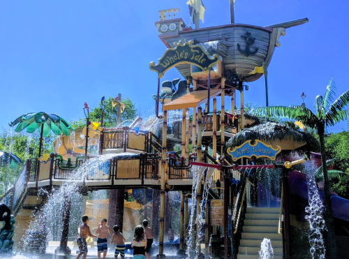 A colorful water park play structure with slides and water features, surrounded by palm trees and children playing.