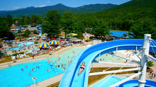 Aerial view of a water park with slides, pools, and people enjoying the sun, surrounded by mountains and trees.