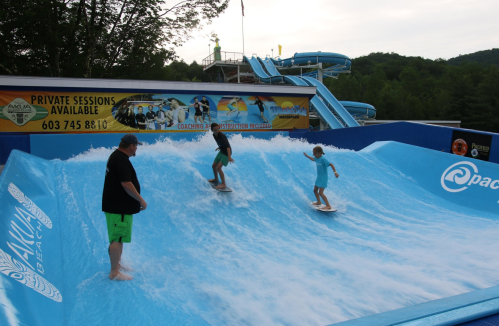 Two children surf on a flow rider while an adult watches nearby, with water slides and a colorful backdrop in the background.