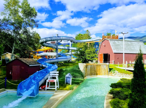 A water park scene featuring a blue water slide, a barn, and a lazy river under a bright sky with fluffy clouds.
