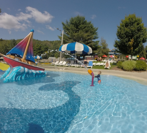A child plays in a pool with a colorful boat sculpture, surrounded by lounge chairs and trees under a blue sky.