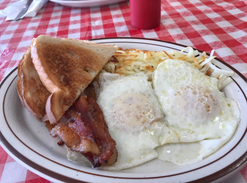 A plate of breakfast featuring two fried eggs, crispy bacon, hash browns, and toasted bread on a checkered tablecloth.