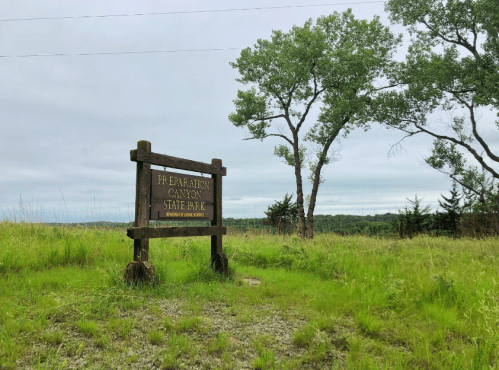 Sign for Preparation Canyon State Park surrounded by green grass and trees under a cloudy sky.