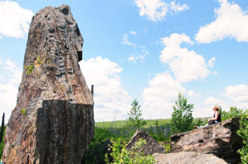 A person sits on a rock, gazing up at a large, towering rock formation under a blue sky with fluffy clouds.