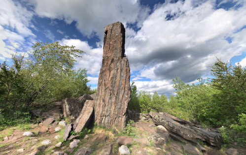A tall, weathered rock formation surrounded by greenery and scattered stones under a cloudy sky.