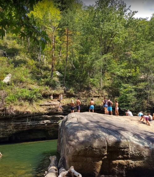 People enjoying a sunny day by a rocky riverbank, some climbing and others relaxing in a lush, green forest setting.