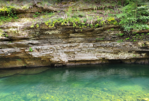 A serene body of water reflecting a rocky, moss-covered cliff surrounded by lush greenery.