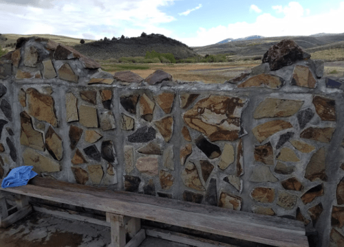 A stone wall with a gap, overlooking a grassy landscape and distant hills under a partly cloudy sky.