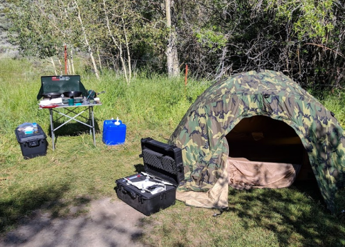 A camouflage tent set up in a grassy area, with camping gear and a table nearby. Trees are in the background.