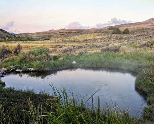 A serene hot spring surrounded by grass and hills, with steam rising gently in a tranquil landscape.