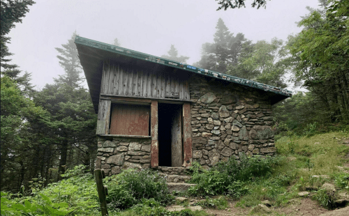 A rustic stone cabin with a slanted roof, surrounded by trees and mist, set in a lush, green forest.