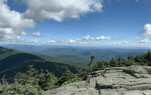 A panoramic view of rolling green mountains under a partly cloudy sky, with rocky foreground and distant peaks.