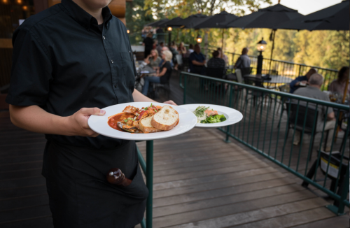 A waiter holds two plates of food while serving on a patio with diners in the background.