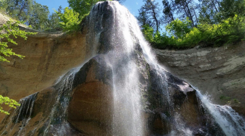 A cascading waterfall flows down a rocky cliff surrounded by lush green trees under a clear blue sky.