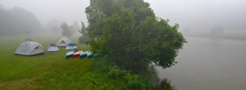Tents set up on a grassy area by a foggy riverbank, with canoes lined up near the water.