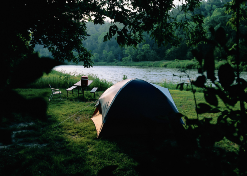 A tent by a river, surrounded by trees, with a table and chairs nearby in a serene camping setting.