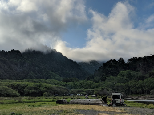 A serene landscape featuring mountains, clouds, and a van near a lush green area with scattered camping spots.