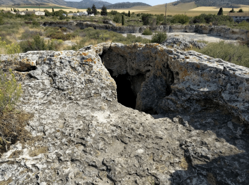 A rocky landscape with a cave entrance, surrounded by sparse vegetation and distant hills under a clear sky.