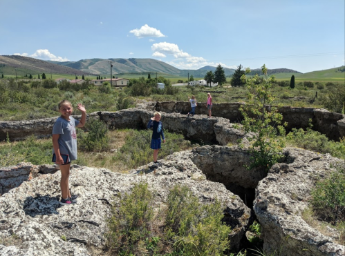 Children wave while standing on rocky terrain with deep holes, surrounded by greenery and hills under a blue sky.