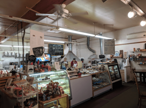 A cozy café interior with a display case of pastries, a kitchen area, and staff preparing food. Warm, inviting atmosphere.
