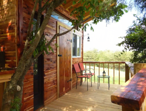 A cozy wooden cabin with a porch, two chairs, and a small table, surrounded by greenery and a peaceful landscape.
