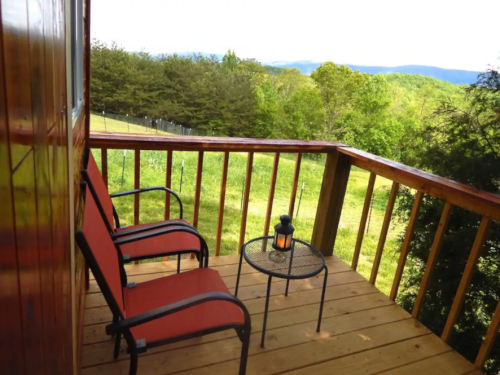 A cozy balcony with two red chairs and a small table, overlooking a lush green landscape and distant mountains.