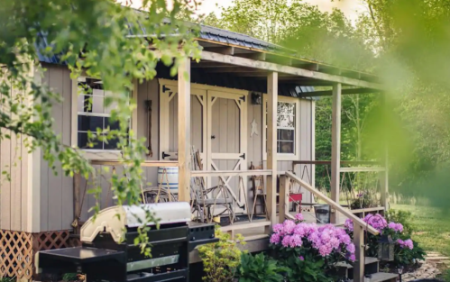 A cozy cabin with a porch, surrounded by greenery and blooming flowers, featuring a grill in the foreground.