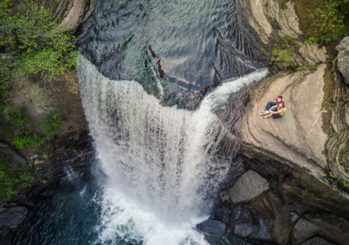 Aerial view of a waterfall cascading into a pool, with two people relaxing on a rock nearby. Lush greenery surrounds the scene.