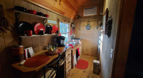 Cozy kitchen with wooden walls, red dishware, a coffee maker, and a small dining area, featuring natural light.