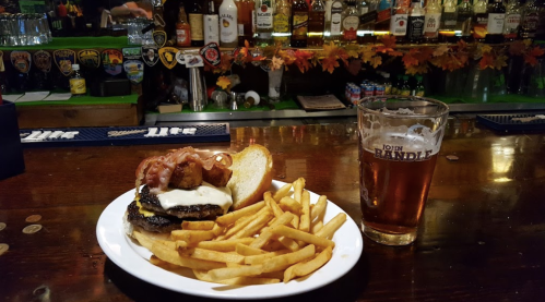 A plate with a burger topped with bacon and cheese, served with fries, alongside a pint of beer at a bar.