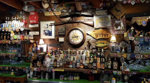 A rustic bar with a variety of liquor bottles, vintage signs, and decorative patches on the walls.