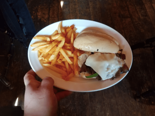A hand holding a plate with a sandwich and fries, featuring a meat patty, cheese, and a soft bun.